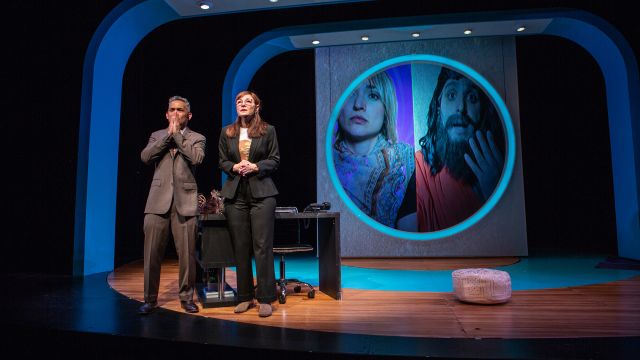 Fred (Ryan Morales) and Joan (Susi Damilano*) hold their breath while Guru Raj (Bobak Cyrus Bakhtiari*, on screen) is being interviewed by Lauren Clark Rose (Ayelet Firstenberg*, on screen)